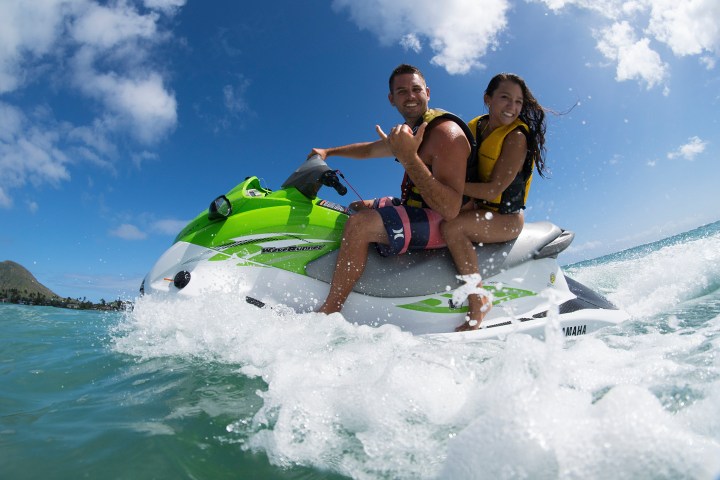 Two people on a jet ski in the ocean, wearing life jackets, with a blue sky and mountain in the background.
