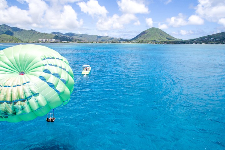 Parasailers over blue ocean near mountain, boat in distance under clear sky.