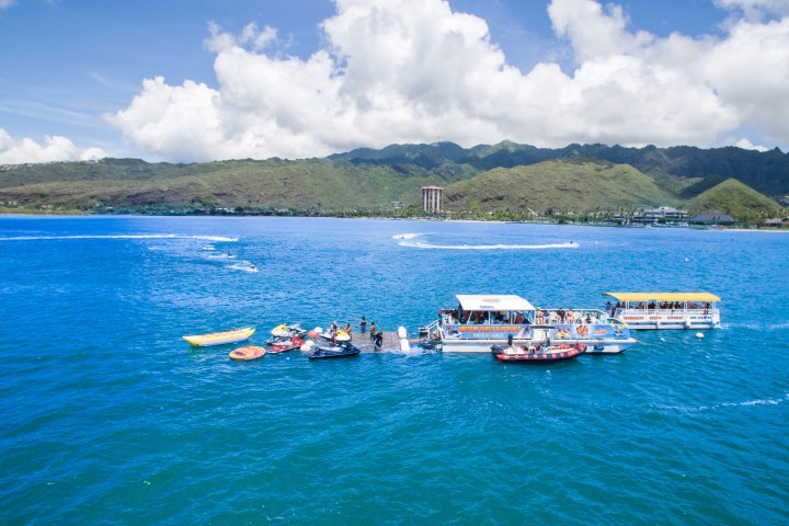 Boats and jet skis on blue water with mountains and clouds in the background.