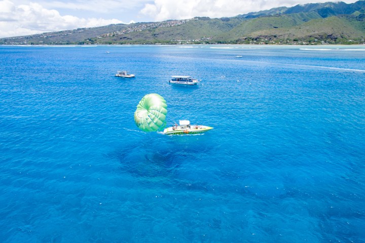 Parasailing boat with green parachute on bright blue ocean, distant shoreline and hills in the background.
