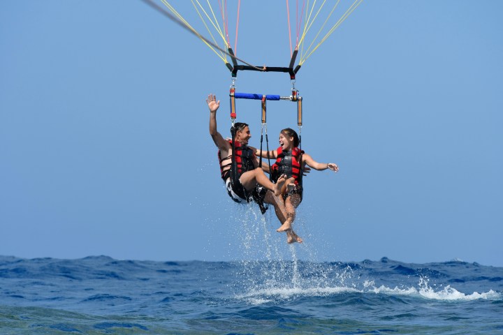 Two people parasailing over water, wearing life vests, smiling and waving, with splashes below.