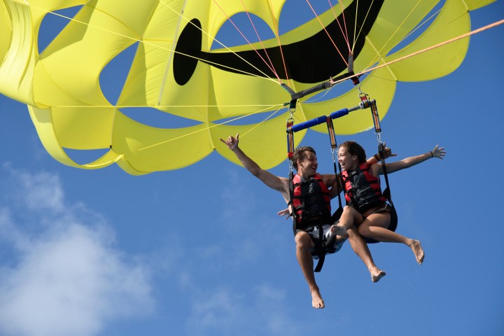 Two people parasailing with a yellow smiley face parachute against a blue sky.